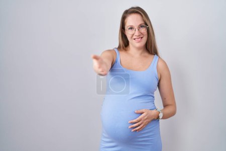 Foto de Mujer embarazada joven de pie sobre fondo blanco sonriendo amistoso ofreciendo apretón de manos como saludo y bienvenida. negocios exitosos. - Imagen libre de derechos