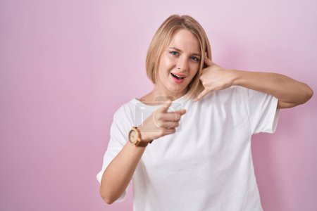 Foto de Young caucasian woman standing over pink background smiling doing talking on the telephone gesture and pointing to you. call me. - Imagen libre de derechos