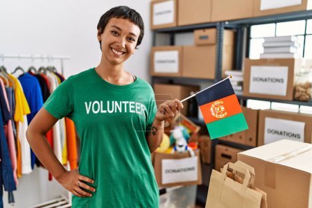 Photo for Young hispanic woman wearing volunteer uniform holding afghanistan flag at charity center - Royalty Free Image