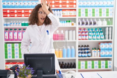 Foto de Hispanic woman with curly hair working at pharmacy drugstore surprised with hand on head for mistake, remember error. forgot, bad memory concept. - Imagen libre de derechos