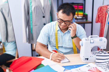 Téléchargez les photos : Jeune homme chinois tailleur parler sur smartphone écriture sur ordinateur portable au magasin de tailleur - en image libre de droit
