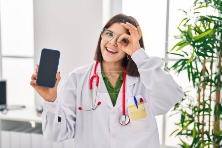 Téléchargez les photos : Jeune médecin hispanique femme montrant écran smartphone souriant heureux faire ok signe avec la main sur les yeux en regardant à travers les doigts - en image libre de droit