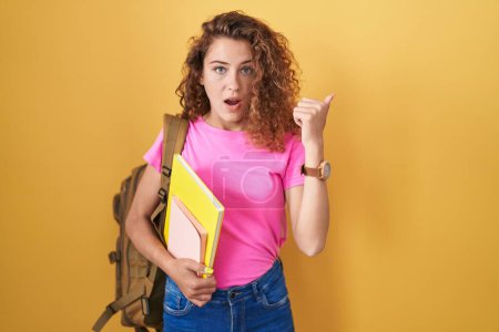 Foto de Young caucasian woman wearing student backpack and holding books surprised pointing with hand finger to the side, open mouth amazed expression. - Imagen libre de derechos