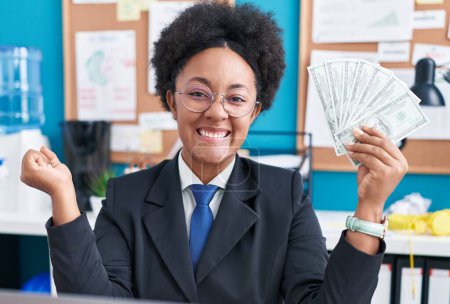 Téléchargez les photos : Belle femme africaine aux cheveux bouclés tenant de l'argent au bureau criant fier, célébrant la victoire et le succès très excité avec le bras levé - en image libre de droit
