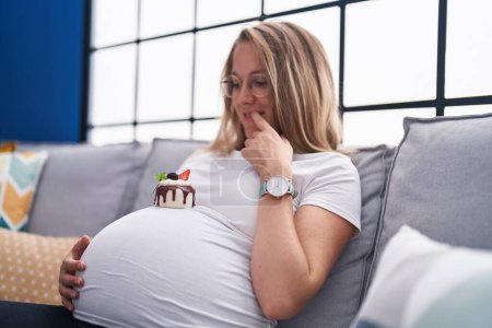 Foto de Joven embarazada buscando pastel tocando vientre en casa - Imagen libre de derechos