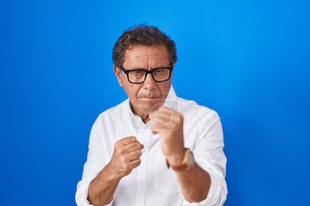 Foto de Middle age hispanic man standing over blue background ready to fight with fist defense gesture, angry and upset face, afraid of problem - Imagen libre de derechos