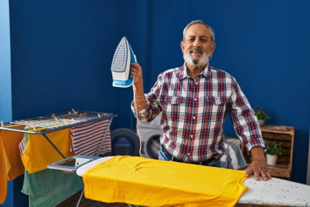Photo for Senior grey-haired man smiling confident holding iron machine at laundry room - Royalty Free Image