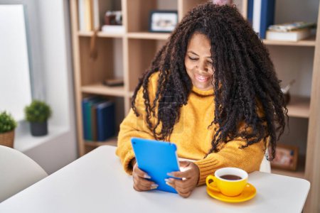 Téléchargez les photos : Femme afro-américaine utilisant touchpad boire du café assis sur la table à la maison - en image libre de droit
