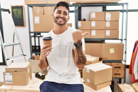 Foto de Young hispanic man working at small business ecommerce drinking a coffee pointing thumb up to the side smiling happy with open mouth - Imagen libre de derechos