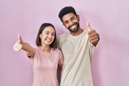 Foto de Joven pareja hispana junta sobre fondo rosa aprobando hacer gesto positivo con la mano, pulgares arriba sonriendo y felices por el éxito. gesto ganador. - Imagen libre de derechos