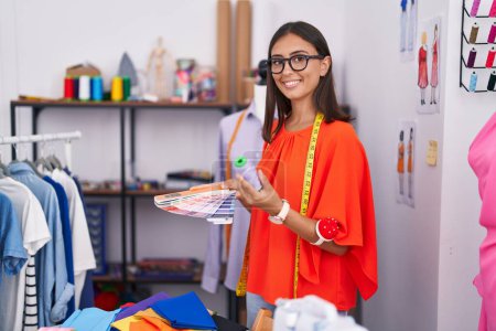 Téléchargez les photos : Jeune belle hispanique femme tailleur souriant confiant tenant t-shirt à la boutique de tailleur - en image libre de droit