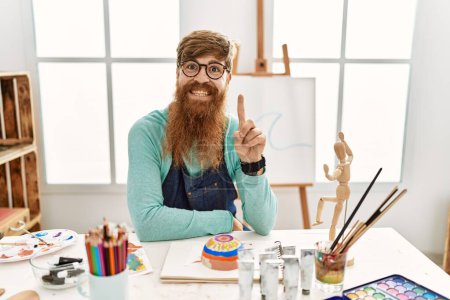 Foto de Redhead man with long beard painting clay bowl at art studio showing and pointing up with finger number one while smiling confident and happy. - Imagen libre de derechos
