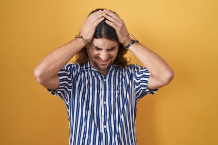 Foto de Hispanic man with long hair standing over yellow background suffering from headache desperate and stressed because pain and migraine. hands on head. - Imagen libre de derechos