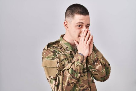 Téléchargez les photos : Young man wearing camouflage army uniform smelling something stinky and disgusting, intolerable smell, holding breath with fingers on nose. bad smell - en image libre de droit