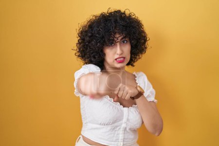 Photo for Young brunette woman with curly hair standing over yellow background punching fist to fight, aggressive and angry attack, threat and violence - Royalty Free Image