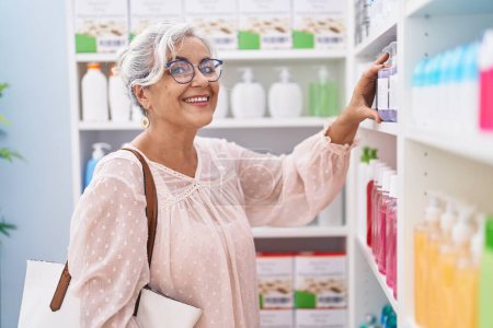 Téléchargez les photos : Femme aux cheveux gris d'âge moyen client regardant étagères à la pharmacie - en image libre de droit