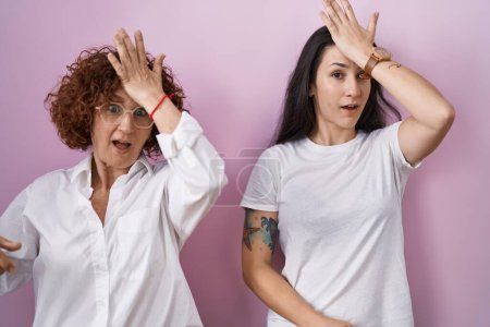 Foto de Hispanic mother and daughter wearing casual white t shirt over pink background surprised with hand on head for mistake, remember error. forgot, bad memory concept. - Imagen libre de derechos