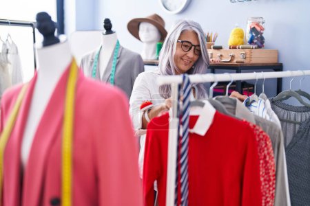 Téléchargez les photos : Femme aux cheveux gris d'âge moyen tailleur souriant confiant tenant des vêtements sur une étagère au magasin de tailleur - en image libre de droit