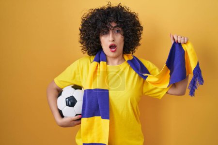Photo for Young brunette woman with curly hair football hooligan holding ball in shock face, looking skeptical and sarcastic, surprised with open mouth - Royalty Free Image