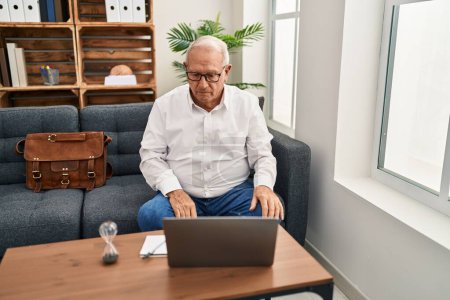 Foto de Senior therapist with grey hair doing online session at consultation office with serious expression on face. simple and natural looking at the camera. - Imagen libre de derechos