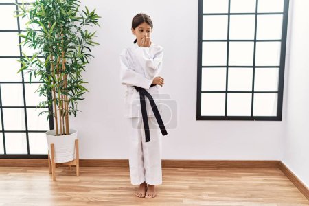 Foto de Young hispanic girl wearing karate kimono and black belt looking stressed and nervous with hands on mouth biting nails. anxiety problem. - Imagen libre de derechos
