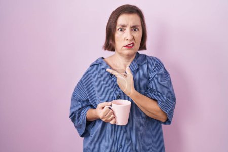 Téléchargez les photos : Middle age hispanic woman drinking a cup coffee pointing aside worried and nervous with forefinger, concerned and surprised expression - en image libre de droit
