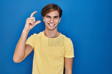Téléchargez les photos : Young man standing over blue background smiling and confident gesturing with hand doing small size sign with fingers looking and the camera. measure concept. - en image libre de droit