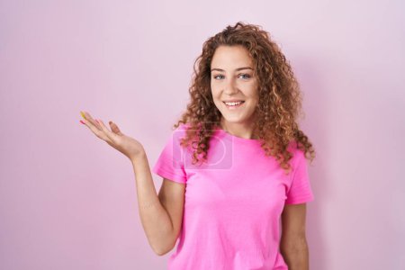 Foto de Joven mujer caucásica de pie sobre fondo rosa sonriente alegre presentando y señalando con la palma de la mano mirando a la cámara. - Imagen libre de derechos
