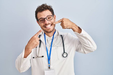 Téléchargez les photos : Jeune homme hispanique portant uniforme de médecin et stéthoscope souriant gai montrant et pointant avec les doigts les dents et la bouche. concept de santé dentaire. - en image libre de droit