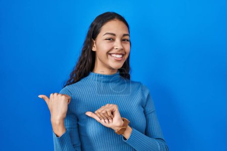 Foto de Young brazilian woman standing over blue isolated background pointing to the back behind with hand and thumbs up, smiling confident - Imagen libre de derechos