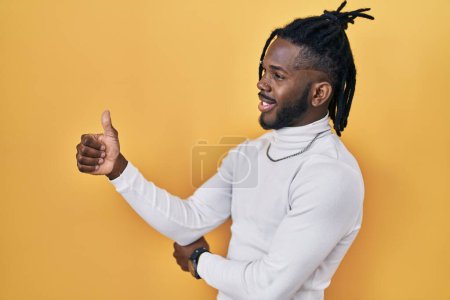 Photo for African man with dreadlocks wearing turtleneck sweater over yellow background looking proud, smiling doing thumbs up gesture to the side - Royalty Free Image