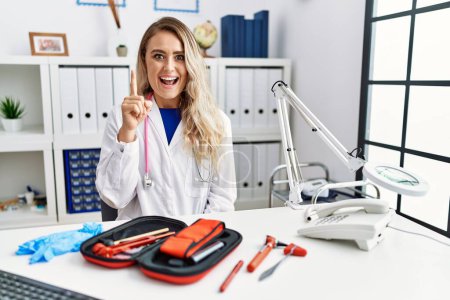 Photo for Young beautiful doctor woman with reflex hammer and medical instruments pointing finger up with successful idea. exited and happy. number one. - Royalty Free Image
