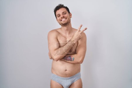 Téléchargez les photos : Young hispanic man standing shirtless wearing underware smiling with happy face winking at the camera doing victory sign with fingers. number two. - en image libre de droit