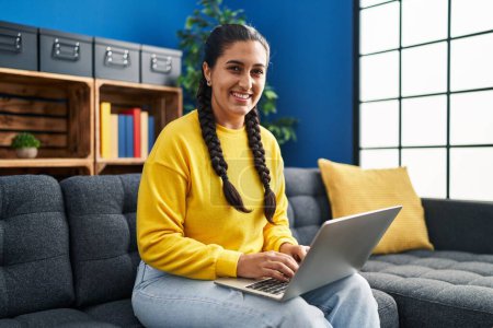 Téléchargez les photos : Jeune femme hispanique utilisant un ordinateur portable assis sur le canapé à la maison - en image libre de droit