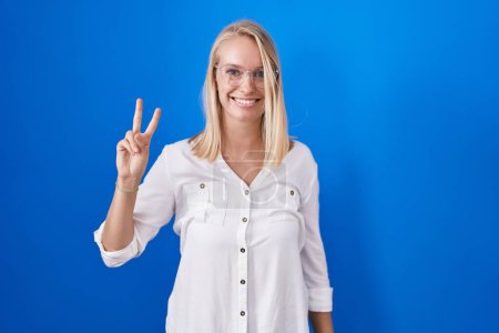 Téléchargez les photos : Jeune femme caucasienne debout sur fond bleu souriant regardant la caméra montrant les doigts faisant signe de victoire. numéro deux. - en image libre de droit
