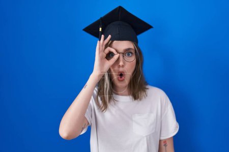 Foto de Blonde caucasian woman wearing graduation cap doing ok gesture shocked with surprised face, eye looking through fingers. unbelieving expression. - Imagen libre de derechos