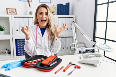 Foto de Young beautiful doctor woman with reflex hammer and medical instruments celebrating surprised and amazed for success with arms raised and open eyes. winner concept. - Imagen libre de derechos