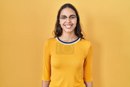 Foto de Young brazilian woman wearing glasses over yellow background looking positive and happy standing and smiling with a confident smile showing teeth - Imagen libre de derechos