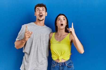 Foto de Young hispanic couple standing together over blue background amazed and surprised looking up and pointing with fingers and raised arms. - Imagen libre de derechos