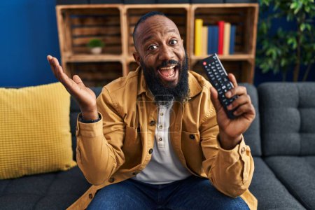 Foto de African american man holding television remote control celebrating victory with happy smile and winner expression with raised hands - Imagen libre de derechos