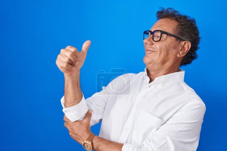Foto de Hombre hispano de mediana edad de pie sobre fondo azul mirando orgulloso, sonriendo haciendo un gesto hacia un lado - Imagen libre de derechos
