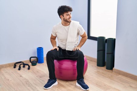 Foto de Hispanic man with beard sitting on pilate balls at yoga room looking away to side with smile on face, natural expression. laughing confident. - Imagen libre de derechos