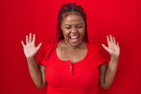 Téléchargez les photos : Femme afro-américaine avec les cheveux tressés debout sur fond rouge célébrant fou et fou de succès avec les bras levés et les yeux fermés hurlant excité. concept gagnant - en image libre de droit