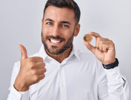 Téléchargez les photos : Handsome hispanic man holding cardano cryptocurrency coin smiling happy and positive, thumb up doing excellent and approval sign - en image libre de droit