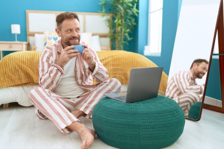 Foto de Middle age man using laptop drinking coffee sitting on floor at bedroom - Imagen libre de derechos