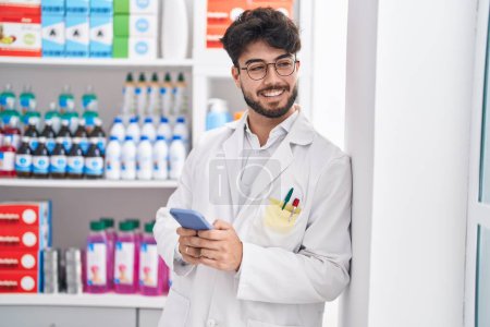 Photo for Young hispanic man pharmacist using smartphone working at pharmacy - Royalty Free Image
