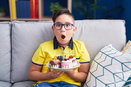 Foto de Joven niño hispano sosteniendo dulces de pastel asustado y sorprendido con expresión sorpresa y asombro, miedo y cara excitada. - Imagen libre de derechos