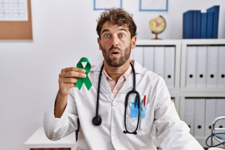 Photo for Young hispanic doctor man holding support green ribbon at clinic scared and amazed with open mouth for surprise, disbelief face - Royalty Free Image