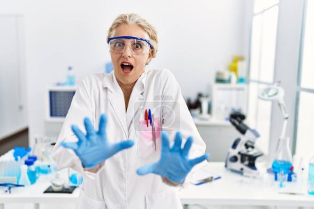 Photo for Middle age blonde woman working at scientist laboratory afraid and terrified with fear expression stop gesture with hands, shouting in shock. panic concept. - Royalty Free Image