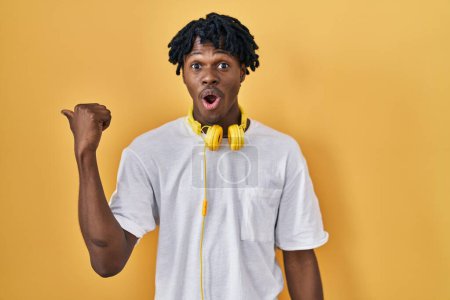 Foto de Young african man with dreadlocks standing over yellow background surprised pointing with hand finger to the side, open mouth amazed expression. - Imagen libre de derechos
